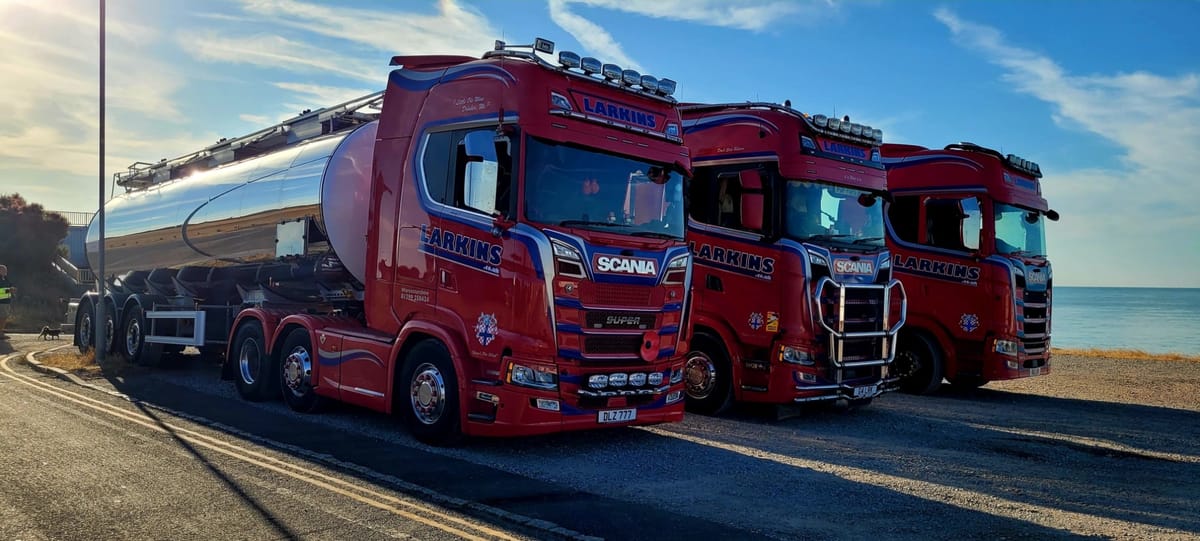Three Larkins Scania tractor units in red livery lined up beside the sea, one coupled to a stainless bulk tanker